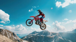 A dynamic shot of a dirt bike rider performing a high jump, capturing the bike mid air with a background of rugged mountains and a clear blue sky