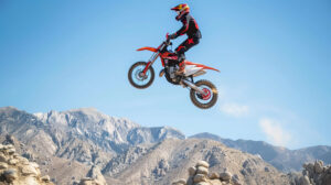 A mesmerizing wallpaper featuring a dirt bike rider in mid air executing a high jump against a backdrop of rugged mountains under a clear blue sky