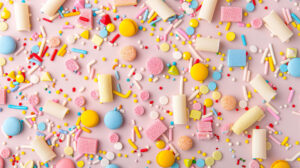 A repeating pattern of various sprinkle shapes and sizes in a rainbow of colors, set against a soft pastel background